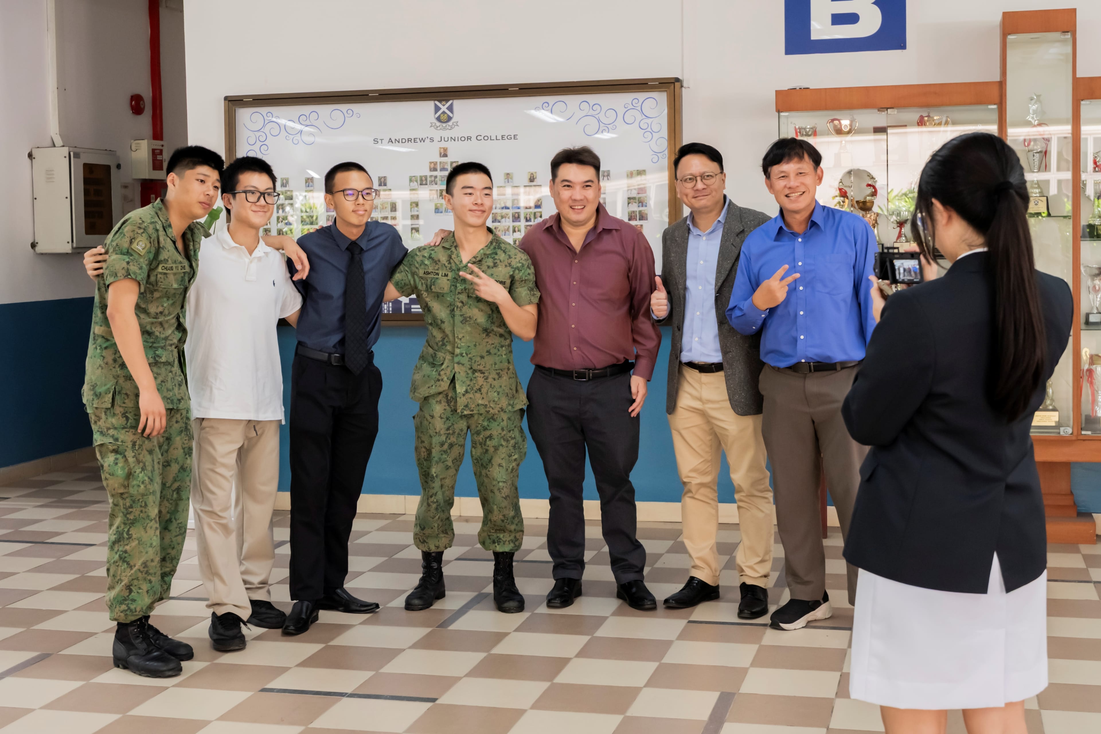 A group consisting of two teachers and 5 students, including two in military camouflage uniforms, pose together while a student councilor takes their picture at St Andrew's Junior College.